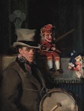 A man dressed in period clothing with a top hat is accompanied by two puppets. One puppet is dressed in a red costume and is holding a stick, while the other is wearing a blue costume with lace details. The scene appears to be dimly lit, suggesting an indoor or theatrical setting.