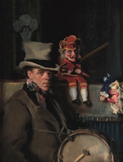 A man dressed in period clothing with a top hat is accompanied by two puppets. One puppet is dressed in a red costume and is holding a stick, while the other is wearing a blue costume with lace details. The scene appears to be dimly lit, suggesting an indoor or theatrical setting.