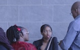 Three people are engaged in conversation against a gray brick wall. The person in the center is facing forward, wearing lipstick, with an attentive expression. On the left, a person is seen from the side with glasses and a red flower in their hair. On the right, a person with glasses is holding a phone and standing slightly turned, also part of the dialogue.