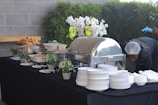 A buffet setup on a black tablecloth features various dishes in chafing dishes and bowls. Decorative elements include potted plants and a floral arrangement with white and yellow flowers. On the side, a person is adjusting or working near the table while wearing gloves and a hairnet. A stack of white plates and bowls is neatly arranged on the right.
