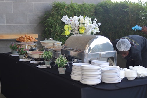 A buffet setup on a black tablecloth features various dishes in chafing dishes and bowls. Decorative elements include potted plants and a floral arrangement with white and yellow flowers. On the side, a person is adjusting or working near the table while wearing gloves and a hairnet. A stack of white plates and bowls is neatly arranged on the right.