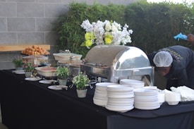 A buffet setup on a black tablecloth features various dishes in chafing dishes and bowls. Decorative elements include potted plants and a floral arrangement with white and yellow flowers. On the side, a person is adjusting or working near the table while wearing gloves and a hairnet. A stack of white plates and bowls is neatly arranged on the right.