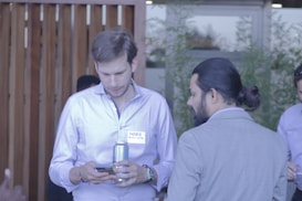 Two men are engaging in conversation outdoors. One man is looking at a phone while holding a can. They are dressed in button-up shirts, and there is a name tag visible on one man. Wooden paneling and greenery are in the background.