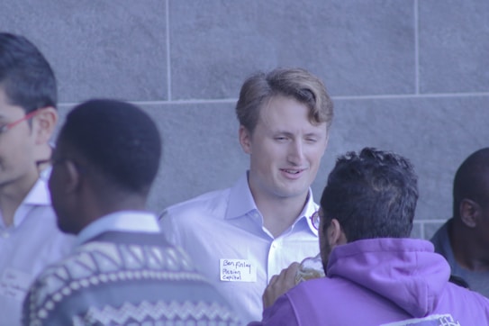 Four men are engaged in conversation, with one of them wearing a name tag. They are in a social or networking setting, possibly at a conference or meetup. The environment suggests a casual or friendly atmosphere.