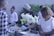 Several people are gathered around a buffet table, with one person serving food onto a plate. They are wearing gloves, indicating a focus on hygiene. The setting includes decorative flowers and a casually dressed group, suggesting a social or communal dining event.
