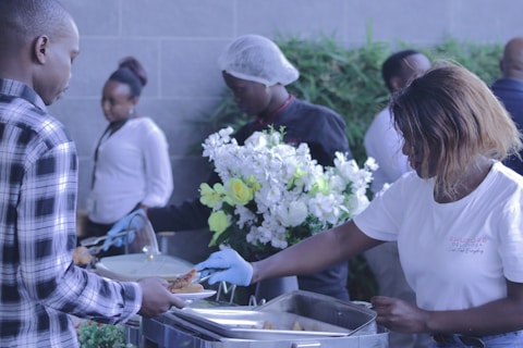 Several people are gathered around a buffet table, with one person serving food onto a plate. They are wearing gloves, indicating a focus on hygiene. The setting includes decorative flowers and a casually dressed group, suggesting a social or communal dining event.