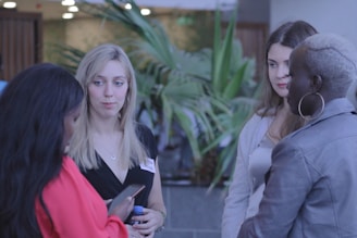 Several women are engaged in conversation, standing in a casual setting with plants in the background. One woman looks at her phone while another listens attentively. They appear to be wearing casual attire and name tags.