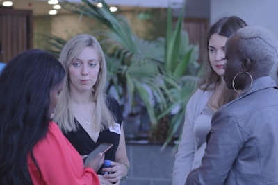 Several women are engaged in conversation, standing in a casual setting with plants in the background. One woman looks at her phone while another listens attentively. They appear to be wearing casual attire and name tags.