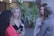 Several women are engaged in conversation, standing in a casual setting with plants in the background. One woman looks at her phone while another listens attentively. They appear to be wearing casual attire and name tags.