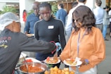 Several people are at a buffet-style food station, being served dishes on plates. One person is wearing a hairnet and serving food, while others are waiting with their plates. The setting appears to be social or professional, with people dressed in casual business attire.