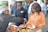 Several people are at a buffet-style food station, being served dishes on plates. One person is wearing a hairnet and serving food, while others are waiting with their plates. The setting appears to be social or professional, with people dressed in casual business attire.