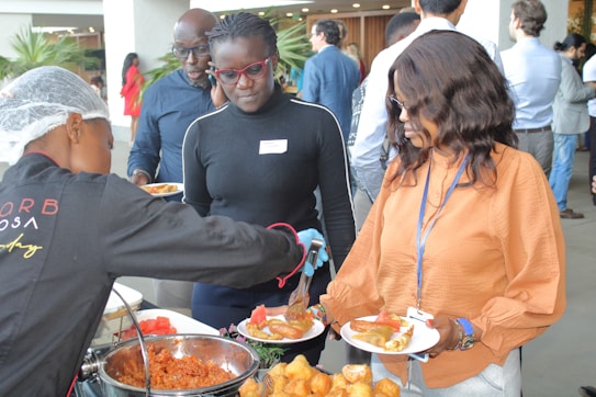 Several people are at a buffet-style food station, being served dishes on plates. One person is wearing a hairnet and serving food, while others are waiting with their plates. The setting appears to be social or professional, with people dressed in casual business attire.
