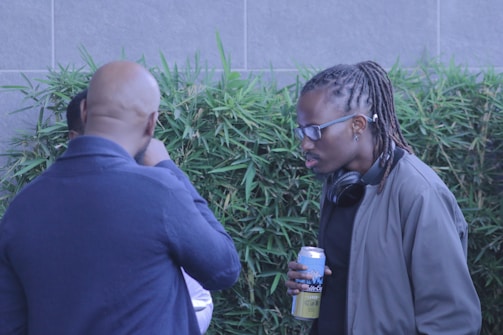Two individuals are in conversation near some green bamboo plants. One person is holding a can and has headphones around their neck. Both appear to be engaged in discussion.