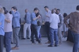 A group of people are gathered in a lobby area, wearing casual and business-casual attire. They are socializing, talking, and some are holding drinks. The background features a modern, tiled wall and a table with refreshments.