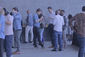 A group of people are gathered in a lobby area, wearing casual and business-casual attire. They are socializing, talking, and some are holding drinks. The background features a modern, tiled wall and a table with refreshments.