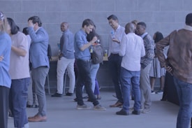 A group of people are gathered in a lobby area, wearing casual and business-casual attire. They are socializing, talking, and some are holding drinks. The background features a modern, tiled wall and a table with refreshments.