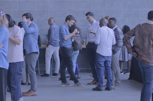 A group of people are gathered in a lobby area, wearing casual and business-casual attire. They are socializing, talking, and some are holding drinks. The background features a modern, tiled wall and a table with refreshments.