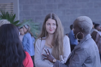A group of people is gathered in a social setting, with a focus on a woman with long hair wearing a light-colored top and another woman with short, dyed hair wearing a leather jacket. They appear to be engaged in conversation, with others in the background creating a lively atmosphere.