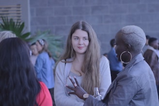 A group of people is gathered in a social setting, with a focus on a woman with long hair wearing a light-colored top and another woman with short, dyed hair wearing a leather jacket. They appear to be engaged in conversation, with others in the background creating a lively atmosphere.