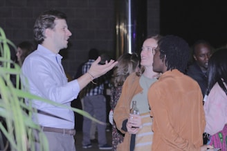 A group of people are gathered together having a conversation in a dimly lit setting. One person is gesturing while others listen attentively. Some guests are holding drinks as they interact.