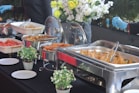 An assortment of buffet-style food dishes arranged on a table, including samosas, fried items, and watermelon. Stainless steel chafing dishes keep the food warm with utensils for serving. Fresh flower arrangements and small potted plants adorn the table, creating an elegant and inviting setup.