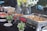 An assortment of buffet-style food dishes arranged on a table, including samosas, fried items, and watermelon. Stainless steel chafing dishes keep the food warm with utensils for serving. Fresh flower arrangements and small potted plants adorn the table, creating an elegant and inviting setup.