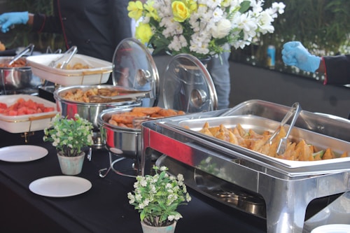 An assortment of buffet-style food dishes arranged on a table, including samosas, fried items, and watermelon. Stainless steel chafing dishes keep the food warm with utensils for serving. Fresh flower arrangements and small potted plants adorn the table, creating an elegant and inviting setup.