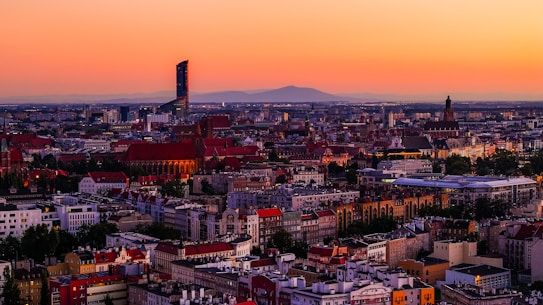 A panoramic view of a city at sunset, highlighting a blend of historic and modern architecture. A tall, modern skyscraper stands prominently, contrasting with the older buildings with red tiled roofs. The sky is dominated by a warm gradient transitioning from orange to pink.