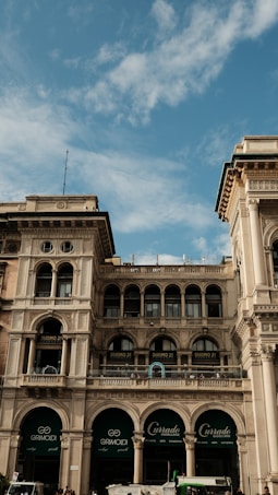 An architectural structure with ornate designs and large arched windows against a backdrop of a partly cloudy blue sky. The building is multi-storied with balconies and decorative columns, featuring signs for various businesses including 'Duomo 21' and 'Grimoldi'.
