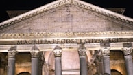 A classical building facade features a large triangular pediment supported by ornate Corinthian columns. The inscription 'MAGRIPPA L F COS TERTIUM FECIT' is prominently displayed across the entablature. The structure is illuminated by warm lights, highlighting its intricate architectural details.