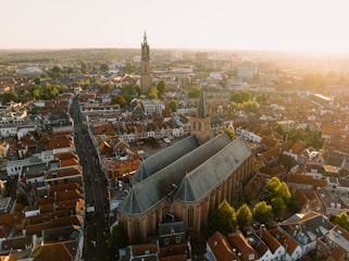 An aerial view of a densely populated urban area with a prominent large church in the center, characterized by its tall spires and grey rooftop. Surrounding the church are numerous buildings and houses with red-tiled roofs. The broader cityscape includes a blend of modern and historical architecture. Streets and trees are visible throughout the city, with a distinctive tower standing out in the background under a warm, golden sunlight.