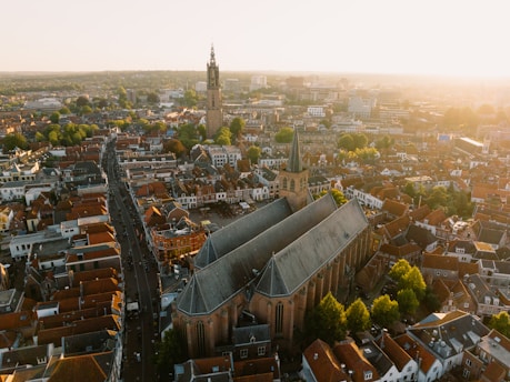 An aerial view of a densely populated urban area with a prominent large church in the center, characterized by its tall spires and grey rooftop. Surrounding the church are numerous buildings and houses with red-tiled roofs. The broader cityscape includes a blend of modern and historical architecture. Streets and trees are visible throughout the city, with a distinctive tower standing out in the background under a warm, golden sunlight.