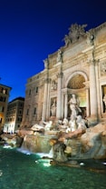 An ornate baroque fountain featuring intricate sculptures and a large classical building facade under a deep blue evening sky. Water cascades elegantly into a pool, surrounded by beautifully carved stone figures.