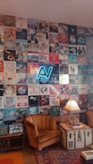 A wall adorned with a collage of vintage music album covers featuring various artists and bands, including a neon sign with letters 'AV' in the center. Below the wall, an old-fashioned room setup includes a brown vintage armchair, a trunk repurposed as a side table with a fringed lamp on top, and a Persian-style rug. The ambient decor suggests a retro, nostalgic atmosphere.