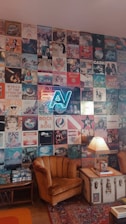 A wall adorned with a collage of vintage music album covers featuring various artists and bands, including a neon sign with letters 'AV' in the center. Below the wall, an old-fashioned room setup includes a brown vintage armchair, a trunk repurposed as a side table with a fringed lamp on top, and a Persian-style rug. The ambient decor suggests a retro, nostalgic atmosphere.