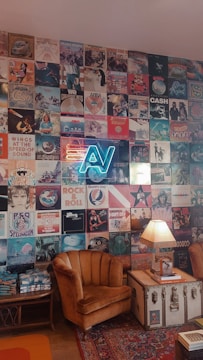 A wall adorned with a collage of vintage music album covers featuring various artists and bands, including a neon sign with letters 'AV' in the center. Below the wall, an old-fashioned room setup includes a brown vintage armchair, a trunk repurposed as a side table with a fringed lamp on top, and a Persian-style rug. The ambient decor suggests a retro, nostalgic atmosphere.