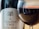 A close-up of a wine glass filled with red wine, reflecting a room with various objects. In the background, a bottle labeled 'Tempus Two Silver Series' features a tree illustration.