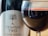 A close-up of a wine glass filled with red wine, reflecting a room with various objects. In the background, a bottle labeled 'Tempus Two Silver Series' features a tree illustration.