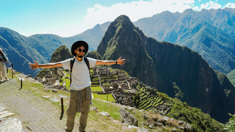 A person with sunglasses and a hat is standing with open arms on a path overlooking the ancient Incan ruins of Machu Picchu. The background has lush green mountains under a clear blue sky.