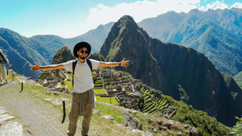 A person with sunglasses and a hat is standing with open arms on a path overlooking the ancient Incan ruins of Machu Picchu. The background has lush green mountains under a clear blue sky.