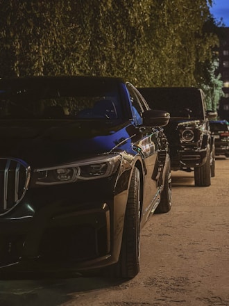 A row of luxury cars parked along a dimly lit street, partially shadowed. The foreground features a sleek black vehicle with chrome detailing, while other high-end vehicles are visible further down the line. The setting suggests an urban environment with a backdrop of trees and buildings.