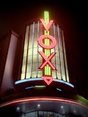 A brightly lit theater marquee prominently featuring the word 'VOX' in vertical neon letters. The sign is illuminated with vibrant red and yellow neon, set against a backdrop of vertical glass panels. The lower part of the image includes a curved awning with additional neon lighting.