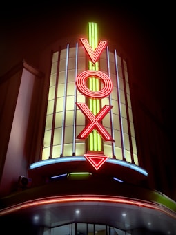 A brightly lit theater marquee prominently featuring the word 'VOX' in vertical neon letters. The sign is illuminated with vibrant red and yellow neon, set against a backdrop of vertical glass panels. The lower part of the image includes a curved awning with additional neon lighting.