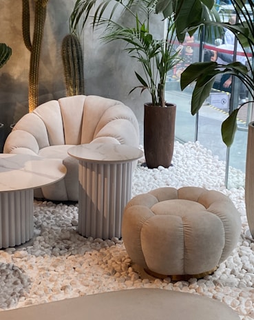 This setting features modern and stylish white furniture, including a plush armchair and a matching round ottoman, both with a tufted design. There are two side tables with marble tops and ribbed cylindrical bases. The space is decorated with potted plants, including tall cacti and leafy plants, placed on a bed of white pebbles. The background includes a large window providing a view of an outdoor industrial space, adding contrast to the interior's cozy and elegant design.