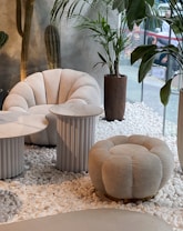 This setting features modern and stylish white furniture, including a plush armchair and a matching round ottoman, both with a tufted design. There are two side tables with marble tops and ribbed cylindrical bases. The space is decorated with potted plants, including tall cacti and leafy plants, placed on a bed of white pebbles. The background includes a large window providing a view of an outdoor industrial space, adding contrast to the interior's cozy and elegant design.