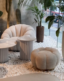 This setting features modern and stylish white furniture, including a plush armchair and a matching round ottoman, both with a tufted design. There are two side tables with marble tops and ribbed cylindrical bases. The space is decorated with potted plants, including tall cacti and leafy plants, placed on a bed of white pebbles. The background includes a large window providing a view of an outdoor industrial space, adding contrast to the interior's cozy and elegant design.