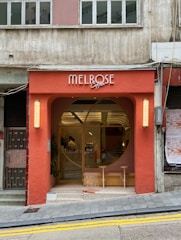 A charming coffee shop with a red exterior facade, featuring a large circular window and the name 'Melrose Coffee' above the entrance. The entrance is adorned with warm lighting and modern decor. The building is positioned on a slight incline and is part of an urban setting.