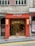 A charming coffee shop with a red exterior facade, featuring a large circular window and the name 'Melrose Coffee' above the entrance. The entrance is adorned with warm lighting and modern decor. The building is positioned on a slight incline and is part of an urban setting.