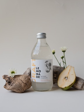 A clear glass bottle with a silver cap, labeled 'Craft Lemonade' showcasing botanic flavors, sits on a light surface. It is surrounded by rustic driftwood, small white flowers, and a sliced pear.