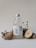 A clear glass bottle with a silver cap, labeled 'Craft Lemonade' showcasing botanic flavors, sits on a light surface. It is surrounded by rustic driftwood, small white flowers, and a sliced pear.
